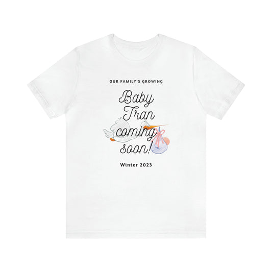 White t-shirt with words: Our Family's Growing. Baby Tran coming soon! Winter 2023 with stork behind the words carrying a baby. Front view.